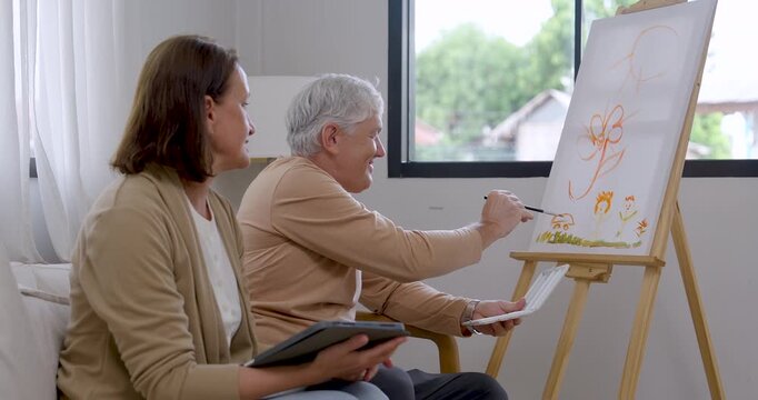 Senior man paints canvas, pauses to review tablet with senior woman, smiles, then resumes brushwork, creating home art therapy activity for emotional wellness, relaxation, bonding, active retirement.