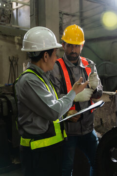 Engineers wearing hardhats and safety vests inspecting metal component and blueprint report in factory.