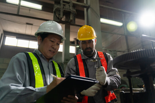 Engineers wearing hardhats and safety vests inspecting metal component and blueprint report in factory.