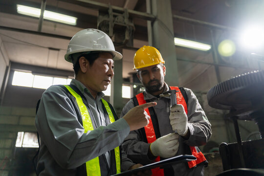 Engineers wearing hardhats and safety vests inspecting metal component and blueprint report in factory.