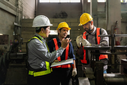 Engineers wearing hardhats and safety vests inspecting metal component and blueprint report in factory.
