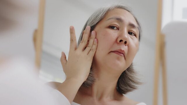 Asian mature woman with natural grey hair applies moisturizing facial serum to her cheek while looking in a mirror. Slow-motion shot.