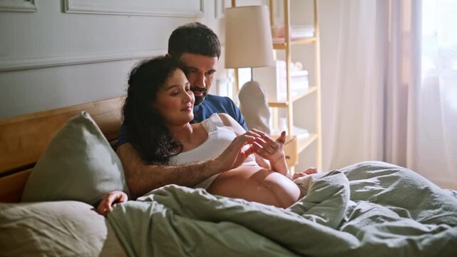 Expecting couple caressing pregnant belly bedroom. Husband touching wife baby