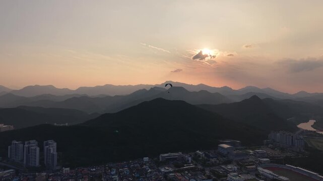 Aerial drone shot orbiting paraglider gliding over mountain landscape with town and river
