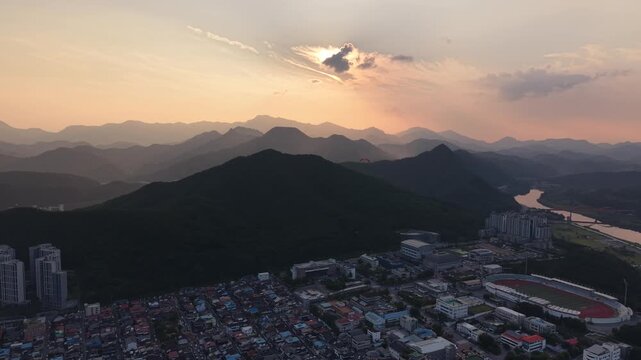 Drone view orbiting over a riverside town and mountain landscape at sunset, distant paraglider in the sky.