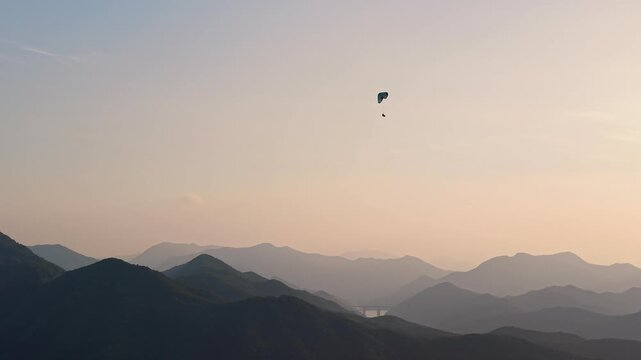 Aerial orbit shot of a paraglider flying over mountain layers during a golden sunset, telephoto lens view.