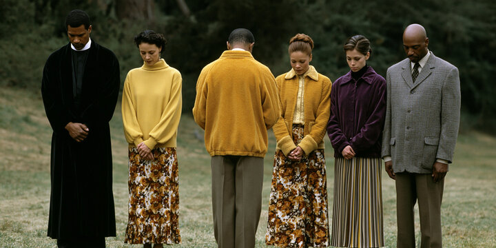 A group of six people standing in a line outdoors with their heads bowed, appearing to be in prayer or deep contemplation