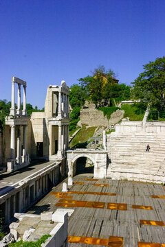 A lone person sits on the stone steps of the ancient Roman Theatre of Philippopolis in Plovdiv, Bulgaria, under a clear blue sky.