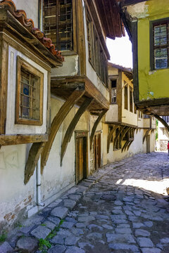 A cobblestone street winds between old, traditional houses with wooden balconies and tiled roofs, bathed in sunlight.