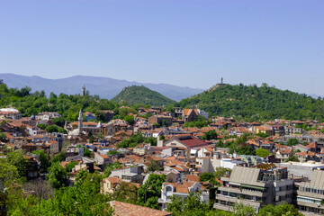Fototapeta premium A panoramic view of the historic city of Plovdiv, Bulgaria, showcasing its distinctive red-tiled roofs, lush green hills, and ancient landmarks under a clear blue sky.