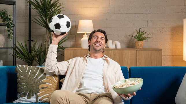 Happy man watching football on sofa, holding soccer ball and bowl of popcorn