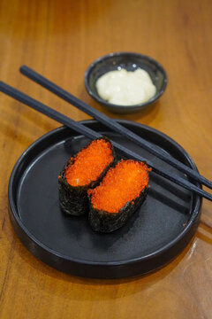 Tobiko gunkan sushi with bright orange flying fish roe wrapped in nori, served on a black plate with chopsticks and mayonnaise at a Japanese restaurant.