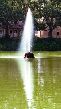 Feuersee in Stuttgart. Vertical photo of a tall water fountain in a city park pond. Urban garden landscape with classical building and trees