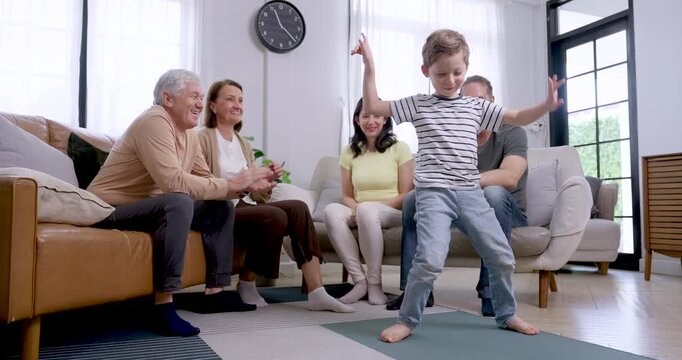 Young son rises from floor, walks toward Caucasian father, and leans across lap as father catches and holds him while Asian mother and senior grandparents smile during family bonding at home.