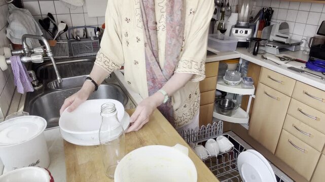 A static kitchen shot showing a woman pushing the upper dishwasher rack inward to access the lower compartment while continuing to load decorative dinner plates.
