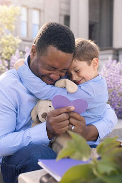 A father hugs his child in a bright outdoors setting, sharing a tender moment with a purple heart card and a stuffed bear, highlighting love, bond, and family.