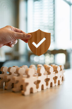 Life insurance and social security concept with hand holding wooden shield with checkmark over group of people figures for employee protection, health safety guarantee and risk management.
