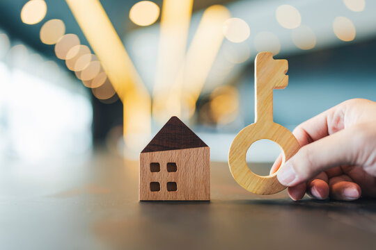 Real estate and property investment concept with hand holding wooden key next to house model on table with warm bokeh background for home loan, mortgage, agent broker and buying new home.