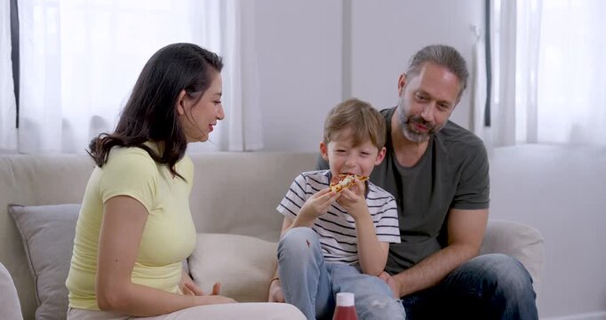 Young son eats pizza, smiles, and gestures while Asian mother and Caucasian father watch and talk on sofa, expressing cheerful family communication, supportive parenting, and warm bonding at home.