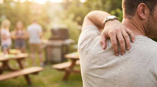 man scratching his itchy back due to an allergic skin reaction or insect bite during an outdoor backyard barbecue picnic with family and friends on a sunny summer afternoon in the garden.
