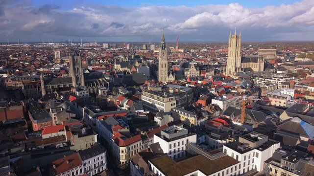 Cinematic aerial drone footage panning over Ghent Belgium reveals stunning medieval cathedral towers and belfry rising above dense historic cityscape with red-roofed buildings under dramatic cloudy.