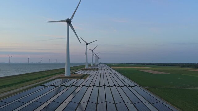 Aerial view of a long row of wind turbines and solar panels along a dutch dike, producing sustainable electricity during a calm sunset