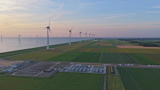 Aerial view of a wind farm with onshore and offshore turbines. Industrial battery storage units and an electrical substation on green fields
