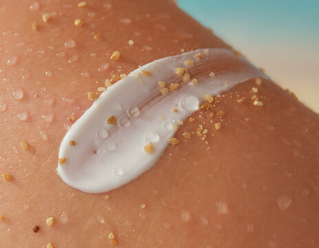 Close-up macro shot of creamy white sunscreen lotion with granular exfoliating particles applied to sun-kissed skin, featuring water droplets and a soft-focus beach background