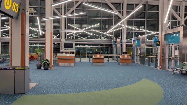 The international airport departures area is empty in the early morning. Bright lights illuminate the space. Empty seating and check-in counters are visible. Few people are moving about.