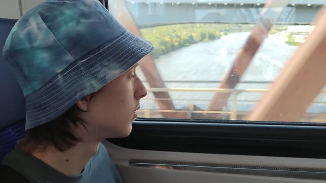 Teenage boy in bucket hat looking out the window while riding train on bridge over river