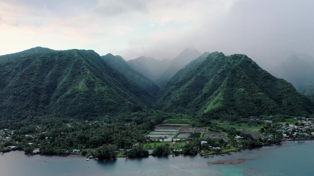 Drone rises slowly over a misty river valley near Teahupoo, Tahiti, revealing the rainforest peaks of Mount Orohena at dawn above a quiet coastal village.
