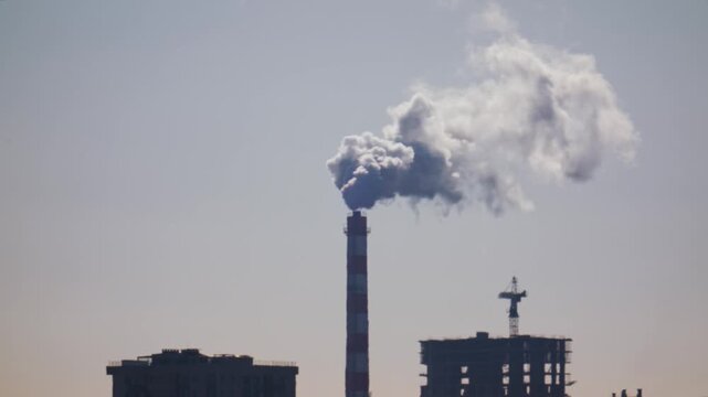 Industrial smoke coming out of tall chimney against clear sky and building site.