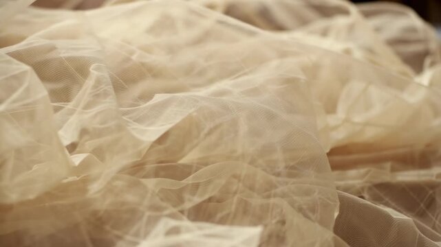 Soft, sheer fabric layers in beige color gently shift and flow, creating a delicate texture in a close-up view with warm lighting and subtle shadows