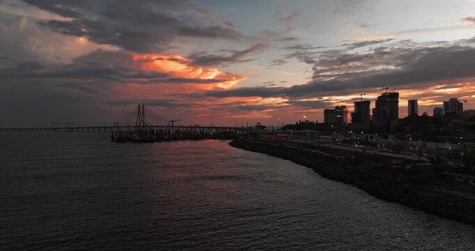 Beautiful sunset view of Mumbai, Maharashtra, with a dramatic orange sky illuminating the city skyline and calm waterfront. The glowing clouds, shimmering water, and striking evening cityscape.