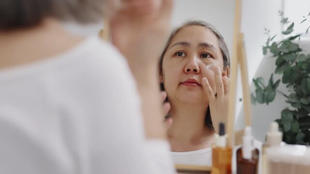 Asian mature woman with natural grey hair applies moisturizing facial cream to her cheek while looking in a mirror. Slow-motion shot.