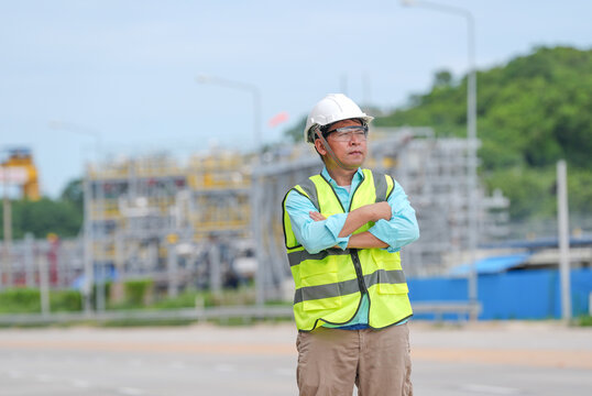 Engineer construction worker helmet safety vest industrial site Asian man standing with crossed arm and serious expression during outdoor inspection near plant equipment road green hill background