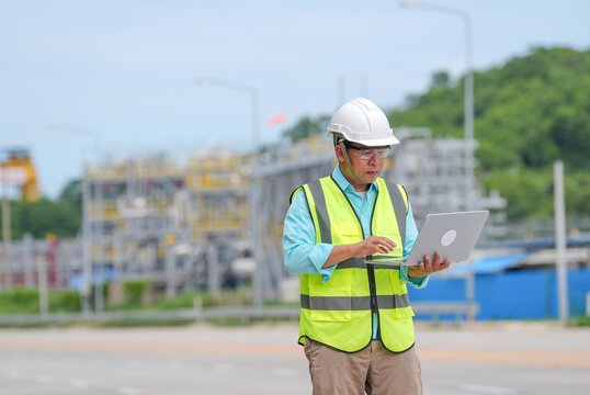 Engineer construction hard hat safety vest laptop industrial outdoor worker using computer at infrastructure site, focused male professional inspecting project under clear sky