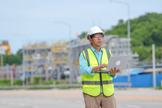 Asian engineer, hard hat, safety vest, laptop, construction site, industrial plant, adult man working outdoors with focused expression near road and green hill background