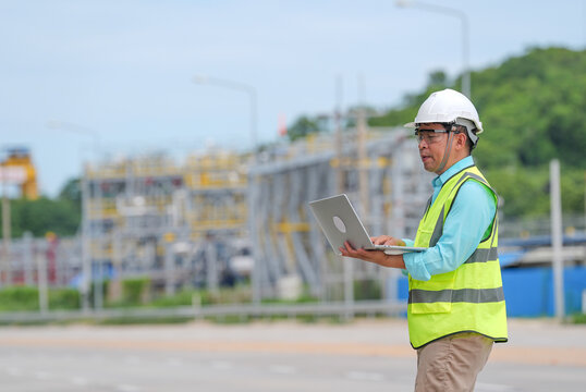 Engineer worker helmet laptop safety vest construction site, focused male professional using computer near industrial facility with road, fence, pipe, hill, and blue sky background