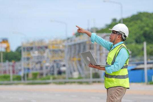 Engineer construction helmet safety vest laptop pointing, male worker giving direction at industrial site with metal structure, road, green hill, blue sky, focused expression