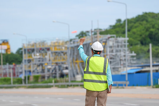 Engineer worker hard hat safety vest industrial plant construction site road inspection back view hand signal at outdoor utility facility with blurred equipment and green hill background