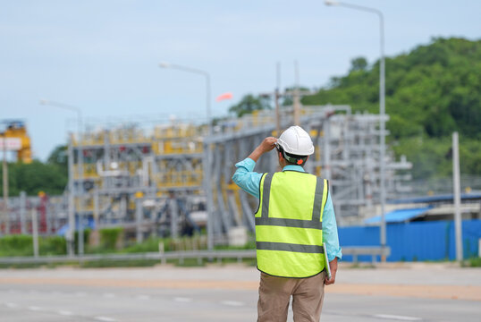 Engineer worker hard hat safety vest industrial plant construction inspection safety male technician surveys utility site near road pipe steel structure green hill