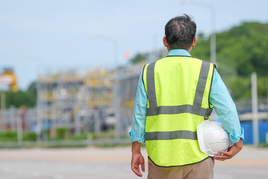 Engineer worker safety vest helmet construction industrial site back view inspection scene with adult man holding hard hat near road and factory area