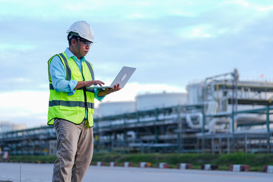 Engineer hardhat safety vest laptop industrial plant refinery worker using computer for inspection maintenance planning and safety monitoring at outdoor energy facility under cloudy sky