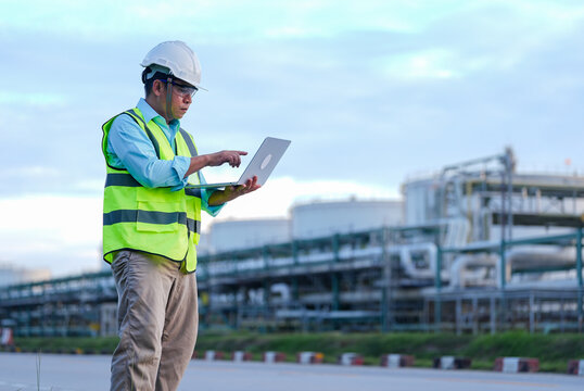 Engineer, hardhat, safety vest, laptop, industrial plant, refinery worker using computer outdoor near pipeline facility with focused expression under cloudy sky