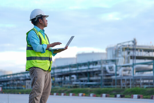 Engineer hard hat laptop safety vest refinery industry inspection technology male worker reviewing data near pipe network at industrial plant under cloudy sky