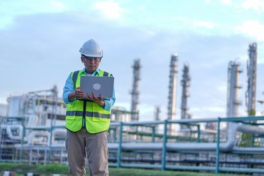 Engineer refinery hardhat laptop safety vest industrial plant pipeline inspection, focused worker checking data during maintenance at petrochemical facility under cloudy sky