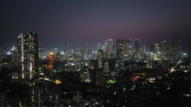 An aerial view of Mumbai's high-rise buildings and beautiful nightlights. The sky is cloudy, and there is medium traffic on the road. The cityscape is filled with modern illuminated buildings