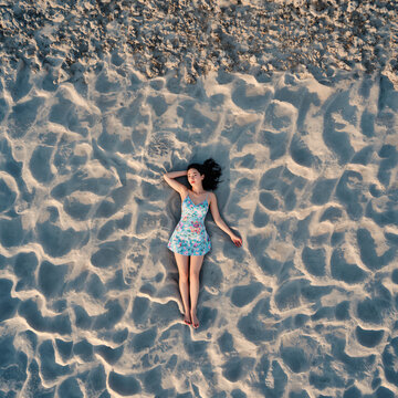 Aerial view of a young woman in a floral dress lying on the sandy beach at sunset.