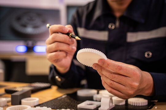 Close-up of hands inspecting a small white plastic gear-like part with a pen at a workbench, with other prototype pieces nearby in a workshop setting